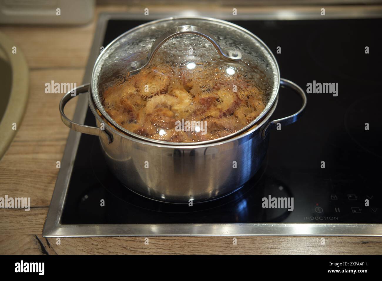 Edelstahl-Topf mit Glasdeckel, der kochende Speisen auf einem Induktionskochfeld enthält. Küchenarbeitsplatte mit Waschbecken im Hintergrund. Stockfoto