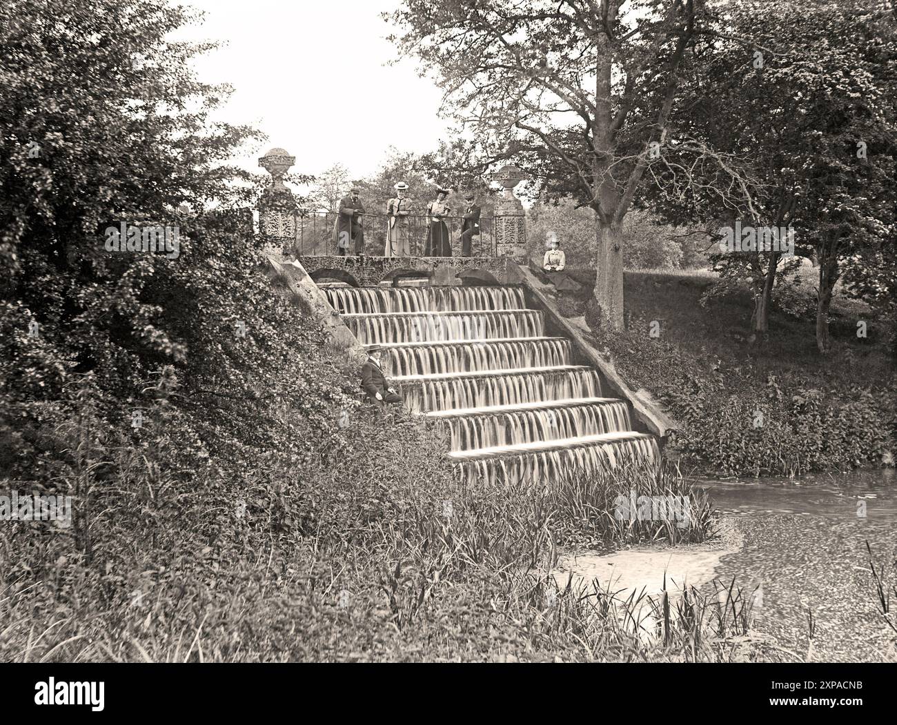 Blick auf die Kaskade im Oriental Garden in den Princess Alexandra Gardens in Leeds Castle, Kent, England, um 1900. Die Kaskade oder der gestufte Wasserfall bildet einen Teil des Flusses Len, der durch das Burggelände fließt. Die modisch gekleideten Viktorianer posieren für die Kamera. Die Kaskade ist noch vorhanden, aber die dekorativen Säulen, das Eisengeländer und die Bogenbrücke existieren nicht mehr. Leeds Castle wurde auf Inseln in einem See errichtet, der vom Fluss Len gebildet wurde. Seit 857 n. Chr. gibt es hier eine Burg – ein viktorianisches und edwardianisches Foto. Stockfoto