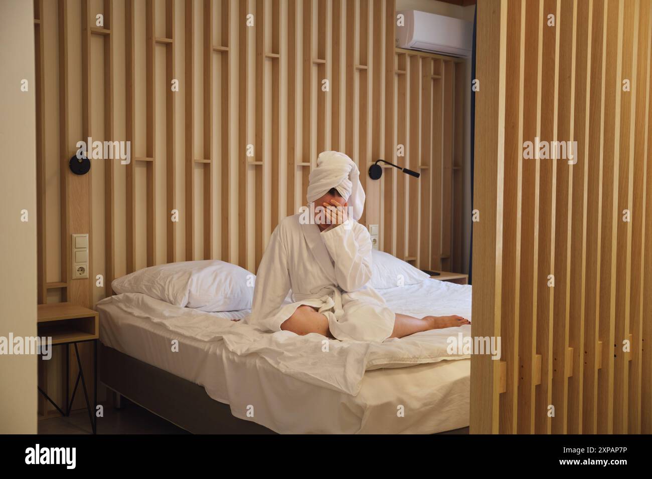 Traurige Frau im weißen Bademantel, die mit dem Kopf in der Hand auf dem Bett sitzt. Stockfoto