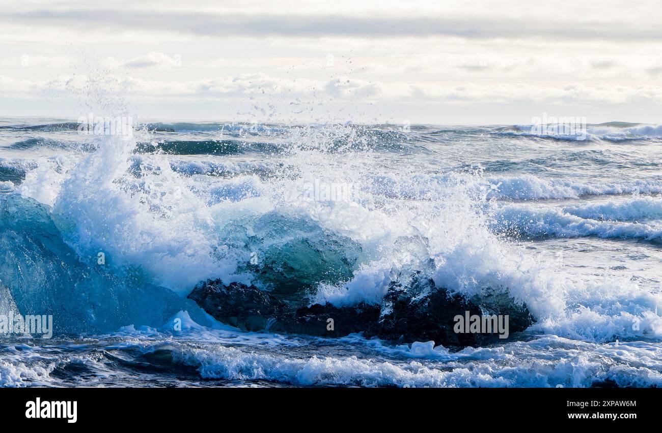Das Meer ist rau und die Wellen stürzen gegen einen Eisberg. Das Wasser ist blau und weiß. Der Himmel ist bewölkt und die Sonne ist nicht sichtbar Stockfoto