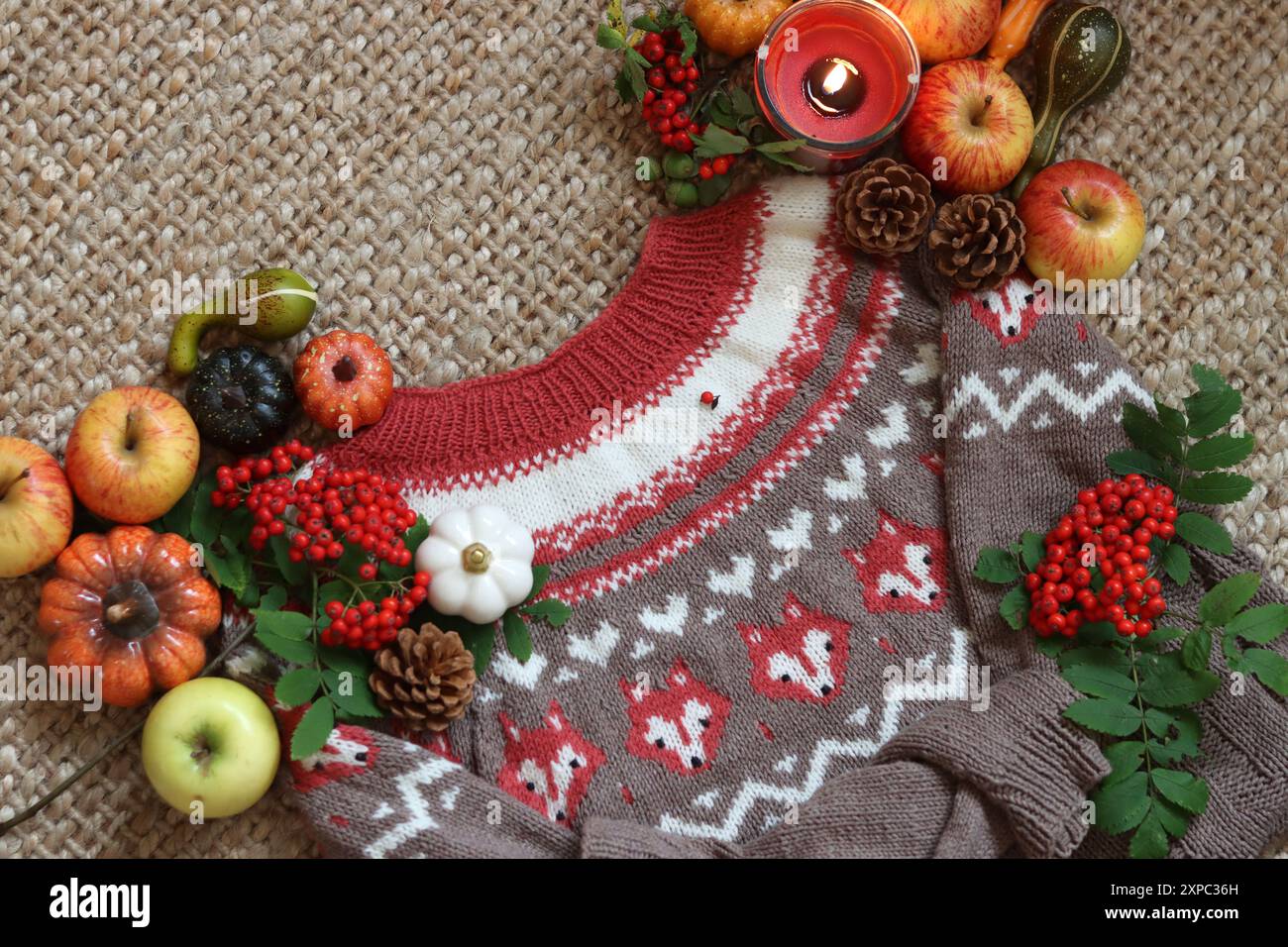 Stillleben im Herbst mit gestricktem braunem und rotem Pullover, Äpfeln, Beeren und Kerze auf Sacktuch Hintergrund. Herbstfüchse Strickmuster Nahaufnahme Foto. Stockfoto