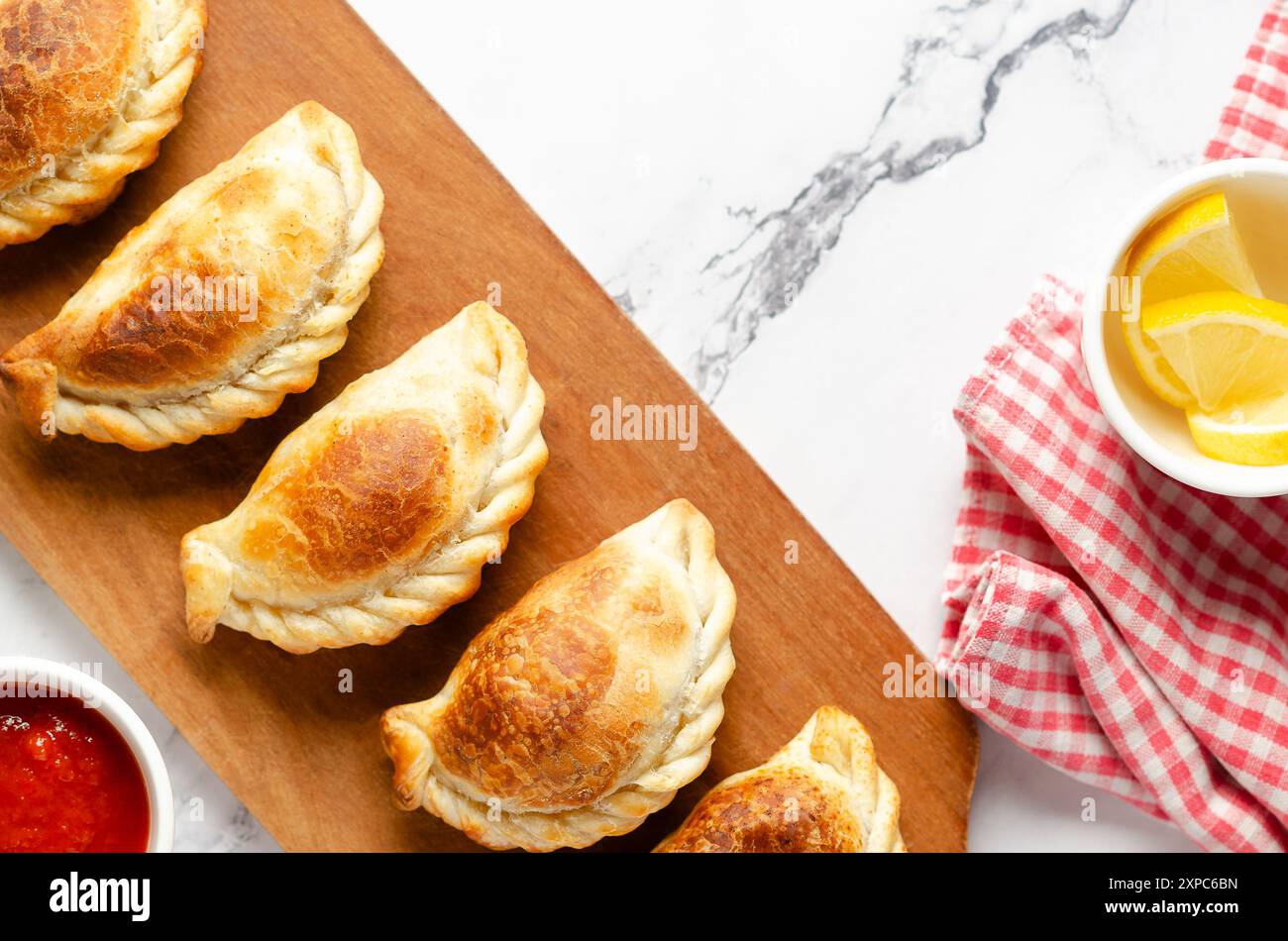 Empanadas aus argentinischem Rindfleisch auf einem Holzbrett, Zitronenscheiben, heiße Sauce und eine rote und weiße Serviette auf weißem Marmorhintergrund. Stockfoto