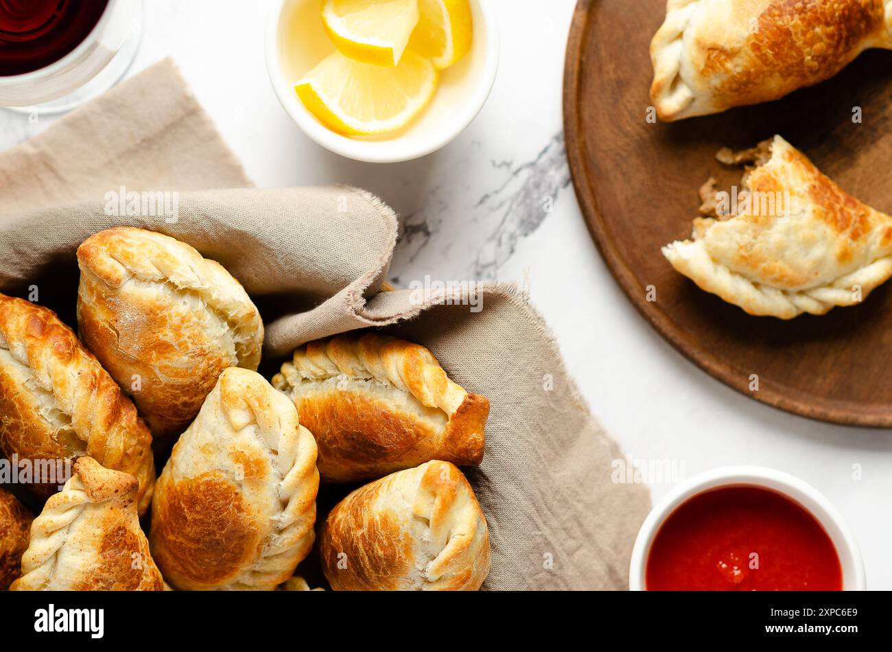 Argentinische Empanadas in einem Korb und auf einem Holzteller, Zitronenscheiben und heiße Soße in weißen Schüsseln auf Marmorhintergrund. Stockfoto