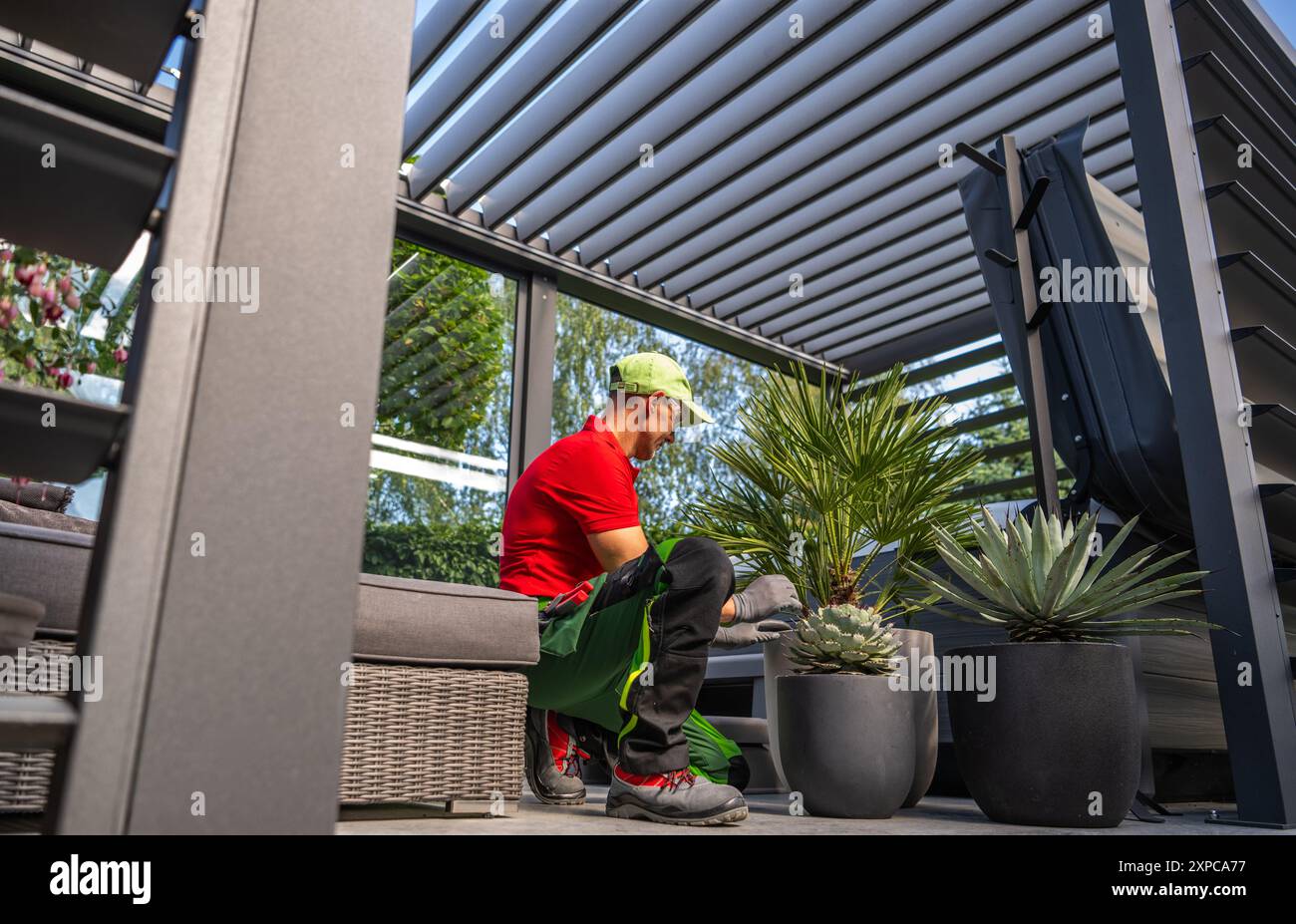 Ein Gärtner in rotem Hemd und roter Mütze kümmert sich um Topfpflanzen auf einer stilvollen Terrasse, umgeben von Grün. Stockfoto