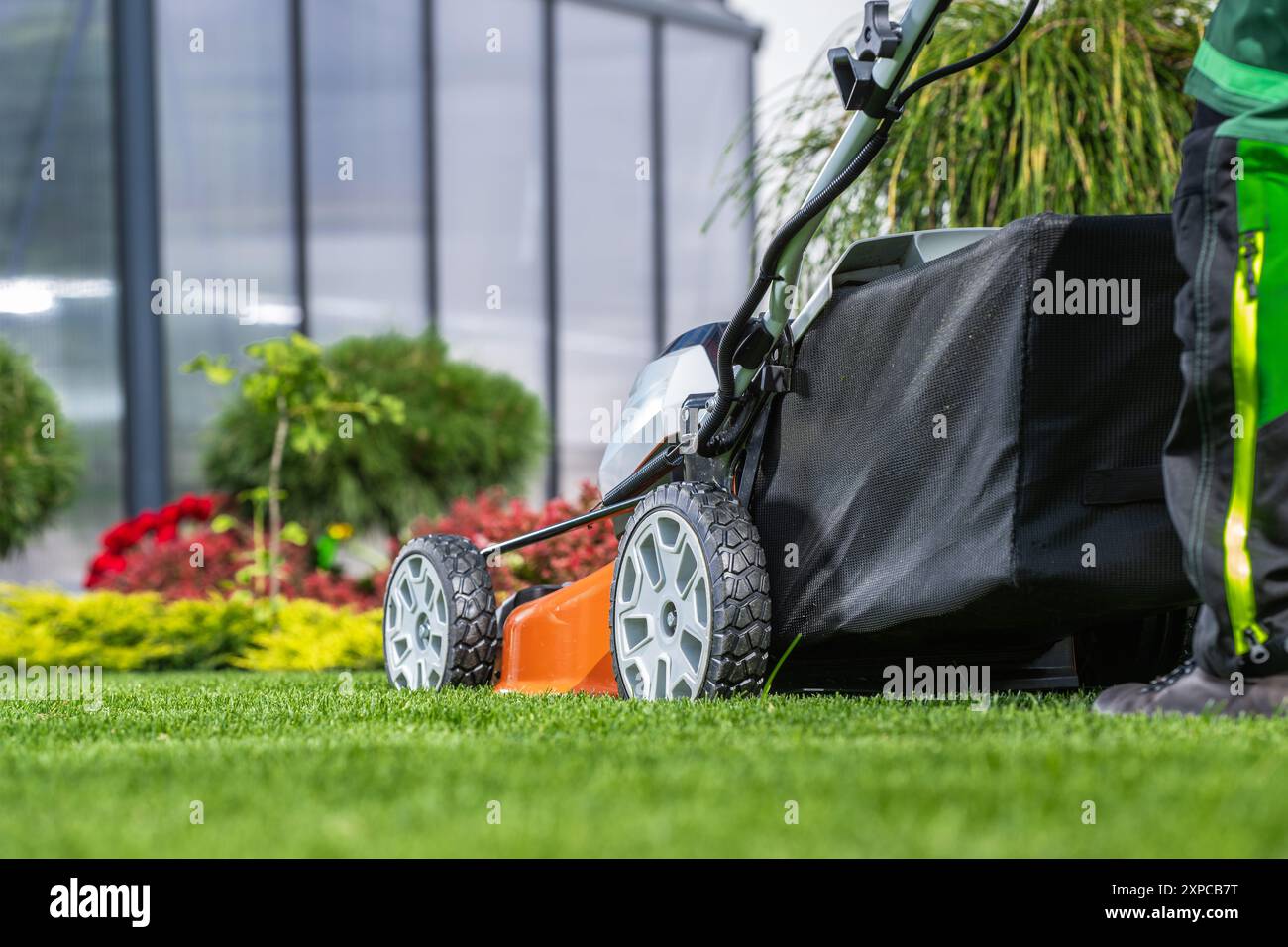 Eine Person mäht einen gepflegten Rasen, umgeben von leuchtenden Blumen, mit einem Rasenmäher, der an einem sonnigen Tag in Betrieb ist. Stockfoto