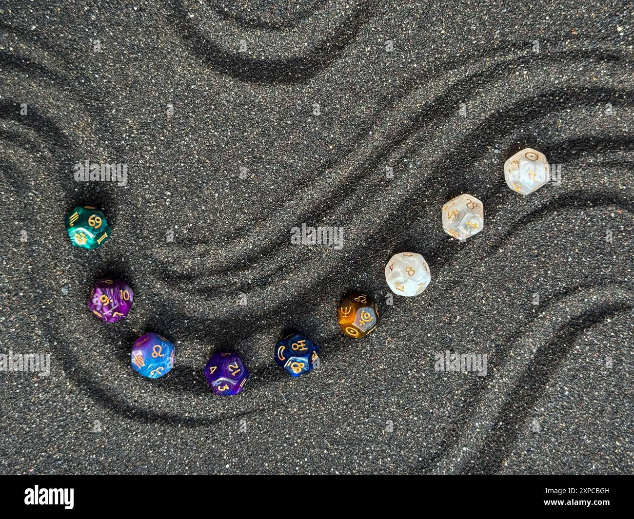 Bunte polyedrische Würfel, die in einer gekrümmten Linie auf einer strukturierten schwarzen Sandoberfläche angeordnet sind Stockfoto