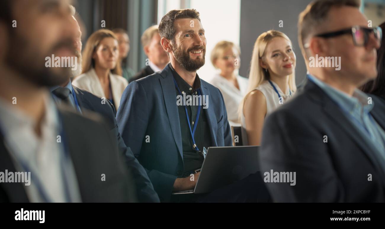 Ein kaukasischer Mann sitzt in einem überfüllten Publikum auf einer IT-Konferenz. Teilnehmer verwenden Laptop-Computer, um Notizen zu machen. Softwareentwickler sehen sich die Präsentation über neue High-Tech-Produkte und Cloud-Lösungen an Stockfoto