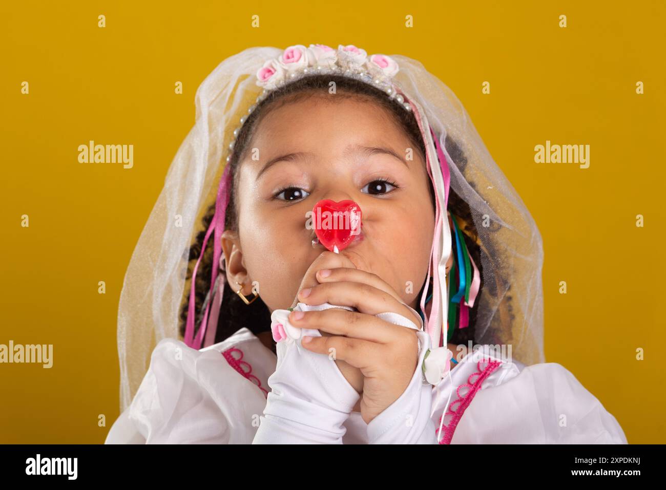 Ein Mädchen als Braut gekleidet, fröhlich und schön, mit einem roten Lutscher im Mund. Porträt mit gelbem Hintergrund im Studio. Stockfoto