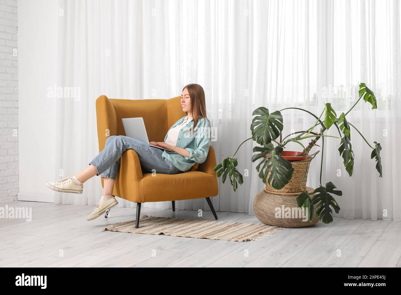 Schöne junge Frau mit Laptop in einem bequemen Sessel in der Nähe des Fensters im Wohnzimmer Stockfoto