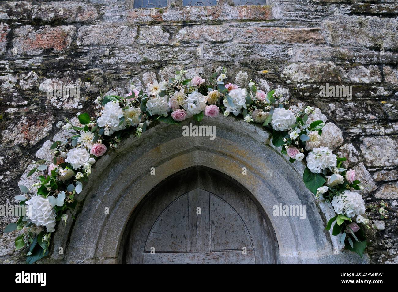 Eine Mischung aus Rosen, Hortensien und Dahlien um den Steinbogen einer hölzernen Kirchentür als Hochzeitsblumen Dekoration, St. Just in Roseland, Cornwall, Großbritannien Stockfoto