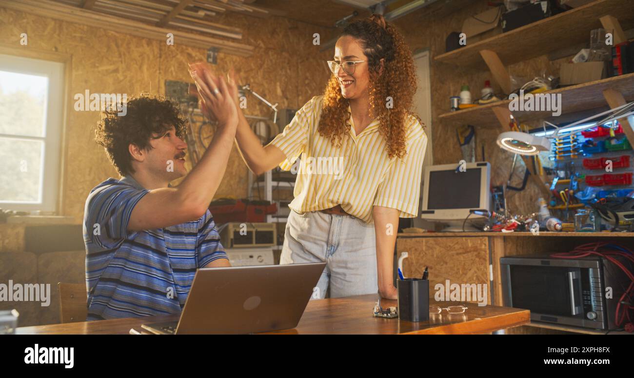 Kaukasischer männlicher Software-Engineer und hispanische weibliche Marketing-Direktorin sprechen und Laptop-Computer in der Garage zu Hause benutzen. Verschiedene Junge Startup-Gründer Arbeiten An Innovativem Online-Service. Stockfoto
