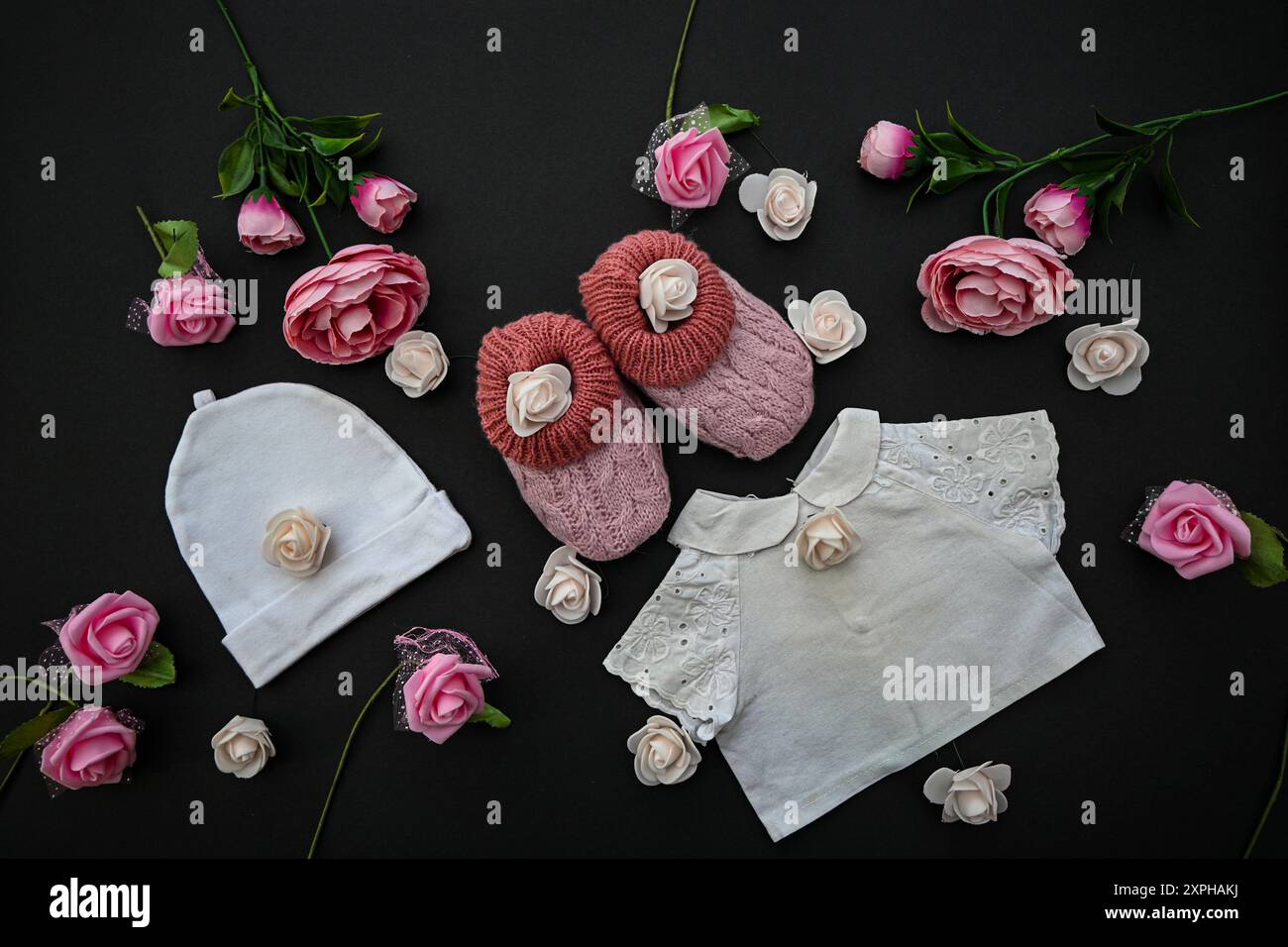 Babykleidung, Schuhe und Rosen auf schwarzem Hintergrund, Babyduschkonzept. Stockfoto