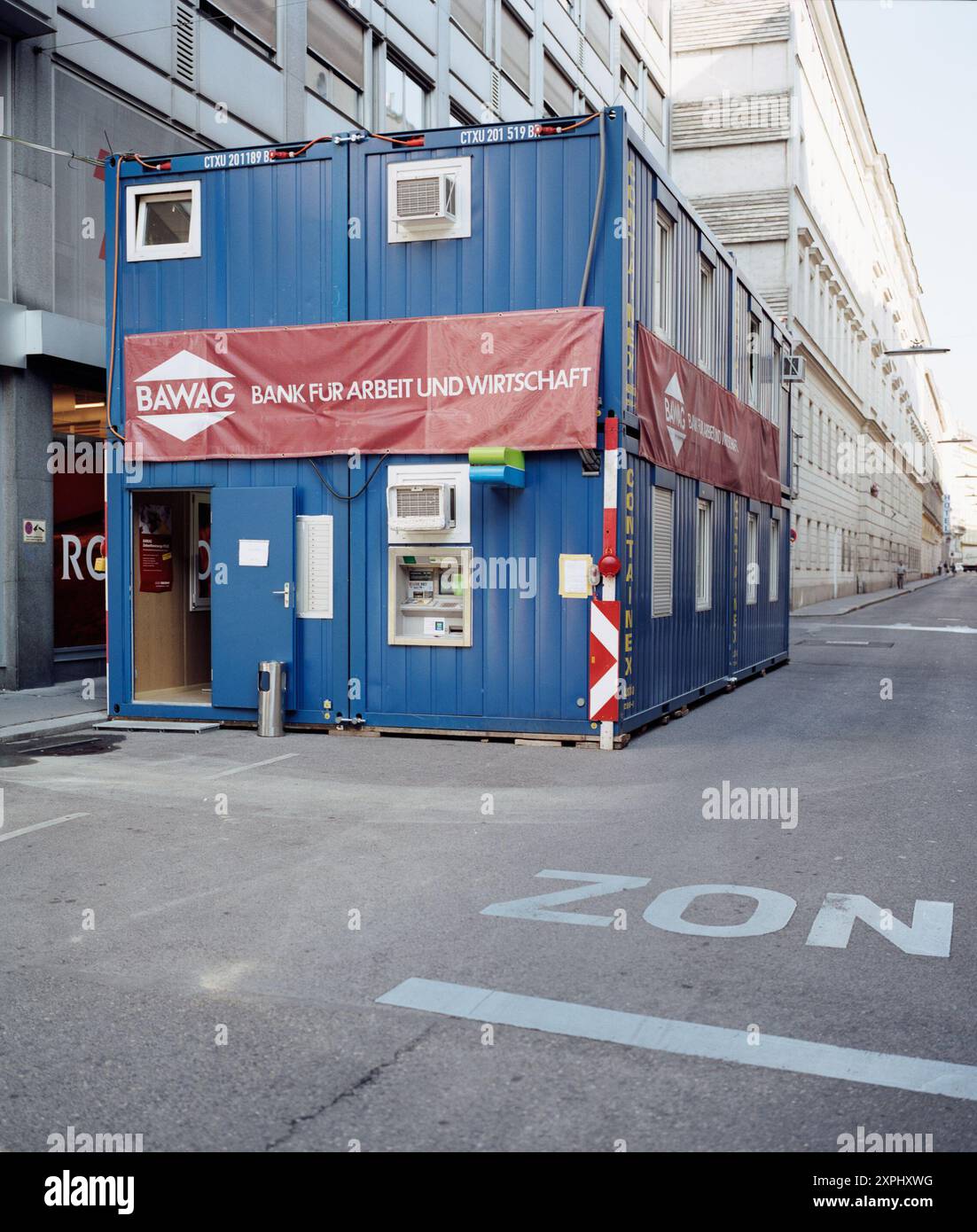 Temporäres Bankbüro in einem blauen Container in einer ruhigen Straße. Die mobile Banking-Einheit ist mit Geldautomaten ausgestattet und mit einem roten Banner markiert, das den Namen der Bank angibt. Stockfoto