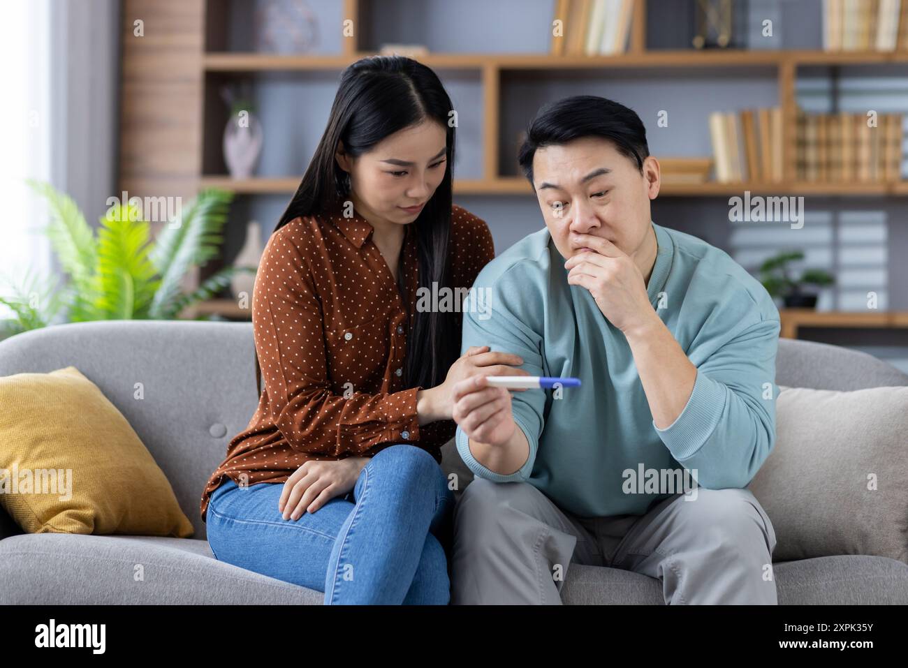Asiatisches Paar sitzt zusammen auf der Couch und untersucht den Schwangerschaftstest. Sie sehen ängstlich und nachdenklich aus und denken über mögliche Lebensveränderungen nach. Regale im Hintergrund mit Pflanzen, Büchern und Dekor. Stockfoto