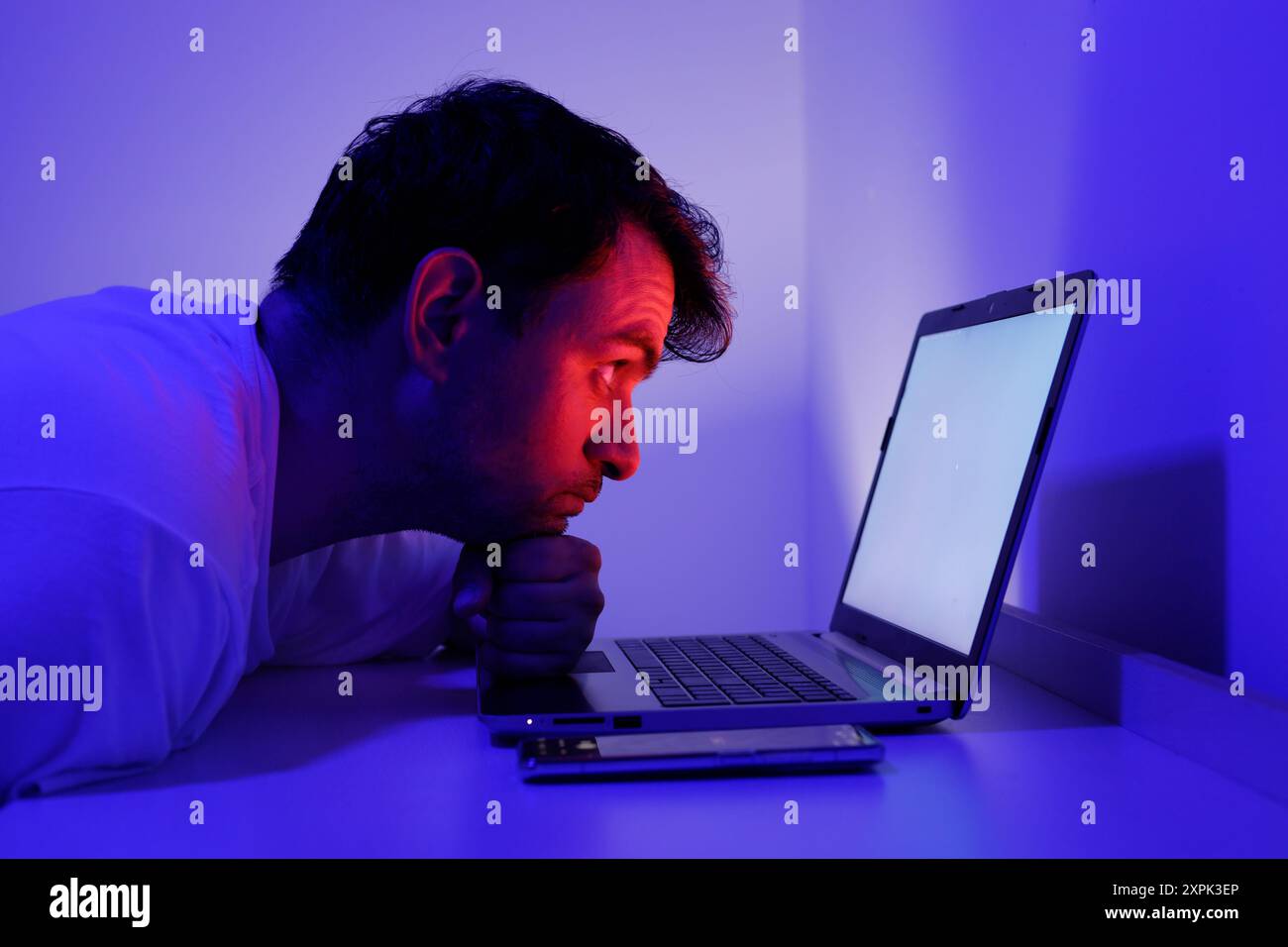 Ein Mann sitzt dicht vor einem Laptop, konzentriert auf den hellen Bildschirm. Seine Stirn ist gefurcht, und er stützt sein Kinn mit seiner Hand. Stockfoto