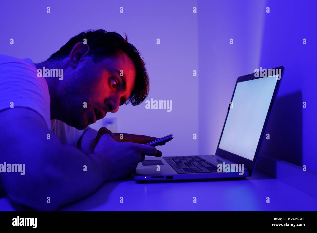 Eine Person lehnt sich aufmerksam nach vorne und starrt auf einen Laptop-Bildschirm, während sie ein Smartphone hält. Die Ambientebeleuchtung ist in einem sanften blauen Farbton gehalten, der ein fokussiertes YE erzeugt Stockfoto