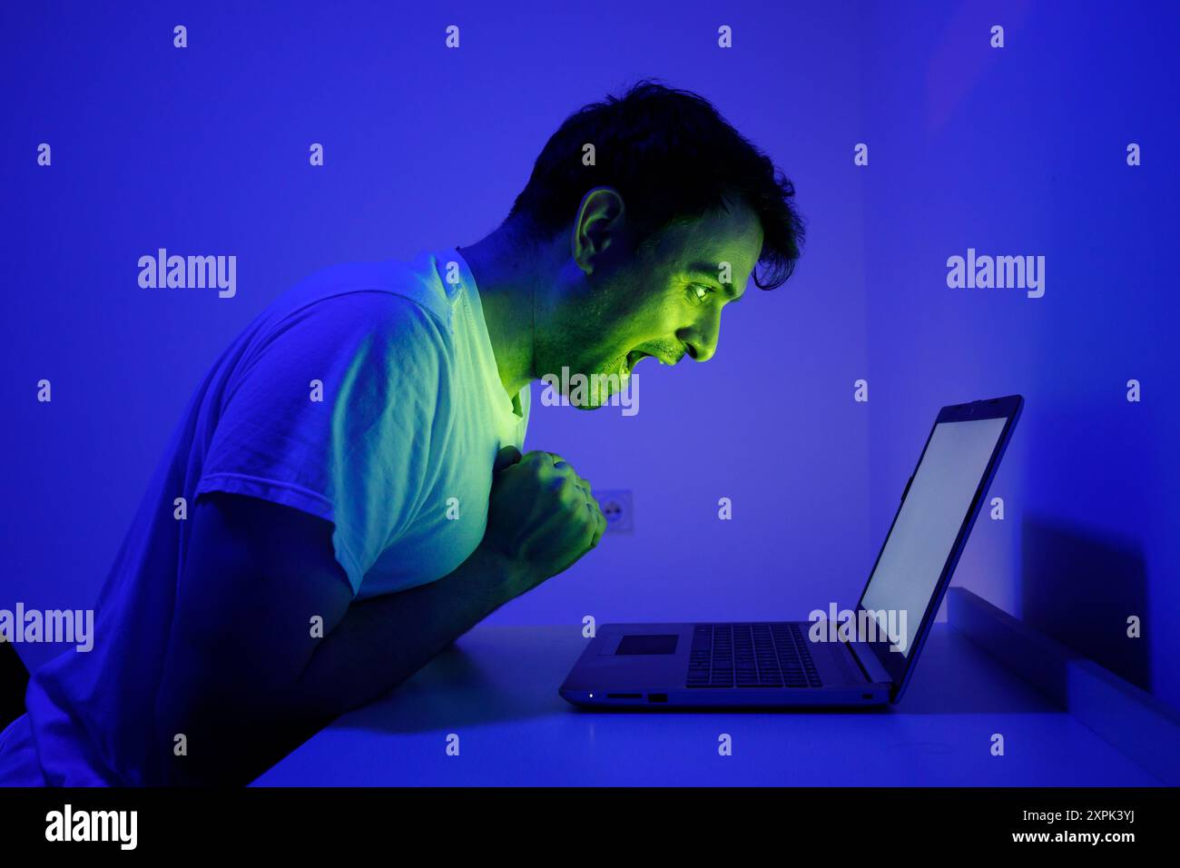 Ein passionierter Gamer sitzt am Schreibtisch und lehnt sich vor Spannung nach vorne, während er auf ein Notebook starrt. Der Raum ist mit einem fesselnden blauen Leuchten beleuchtet, en Stockfoto