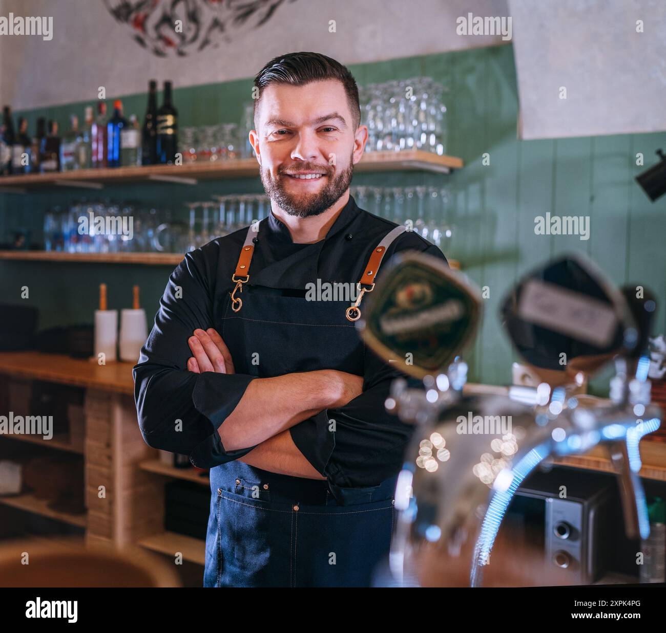 Porträt eines fröhlich lächelnden bärtigen Barkepers in schwarzer Uniform mit Schürze am Bartheken mit gezapftem Bier. Erfolgreiche Mitarbeiter, harte Arbeit, Stockfoto