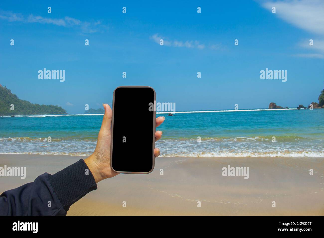 Smartphone-Mockup am Meeresstrand, Nahaufnahme einer weiblichen Hand, die ein Mobiltelefon mit leerem schwarzen Bildschirm als Kopierraum an der Küste im Sommer hält, se Stockfoto
