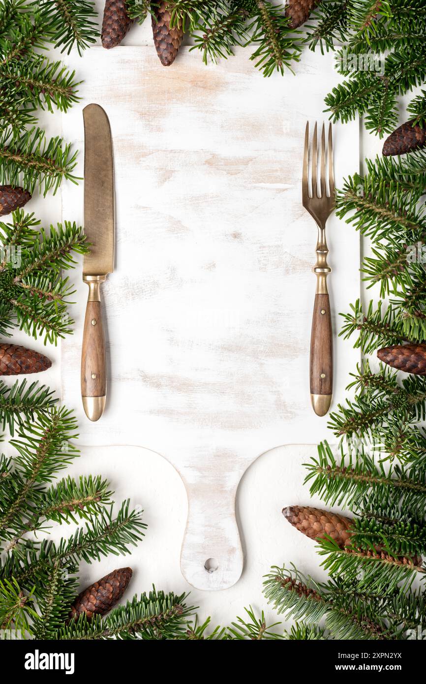 Hintergrund des Weihnachtsmenüs mit weißem Holzschneidebrett, Messer, Gabel und frischen Tannenzweigen mit Kegeln. Weihnachten oder Neujahr Hintergrund. Kreatives Mockup für Winterferien Stockfoto