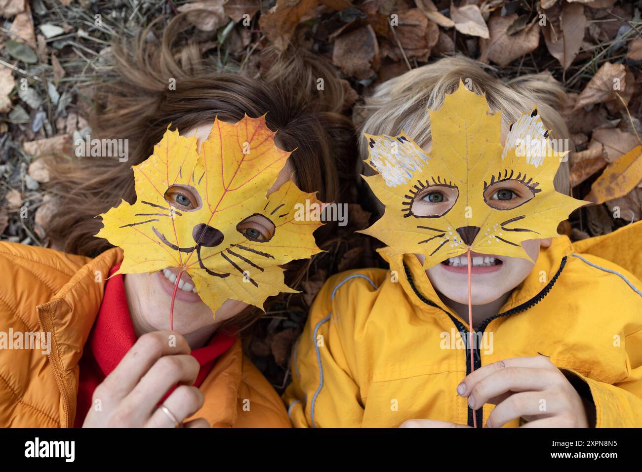 Frau und Kind verstecken ihre Gesichter hinter herbstlichen Ahornblättern, auf denen das Gesicht einer Katze geschnitzt ist. Spaß in der Natur, positives Denken. Familienfreude, happ Stockfoto