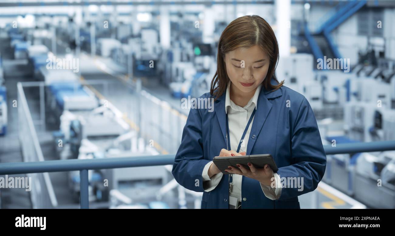 Junge asiatische Ingenieurin, die auf einer Plattform steht und einen Tablet-Computer in einer Elektronikfabrik benutzt. Maschinen werden gewartet, Spezialist überwacht den Fortschritt mithilfe der Online-Software Stockfoto