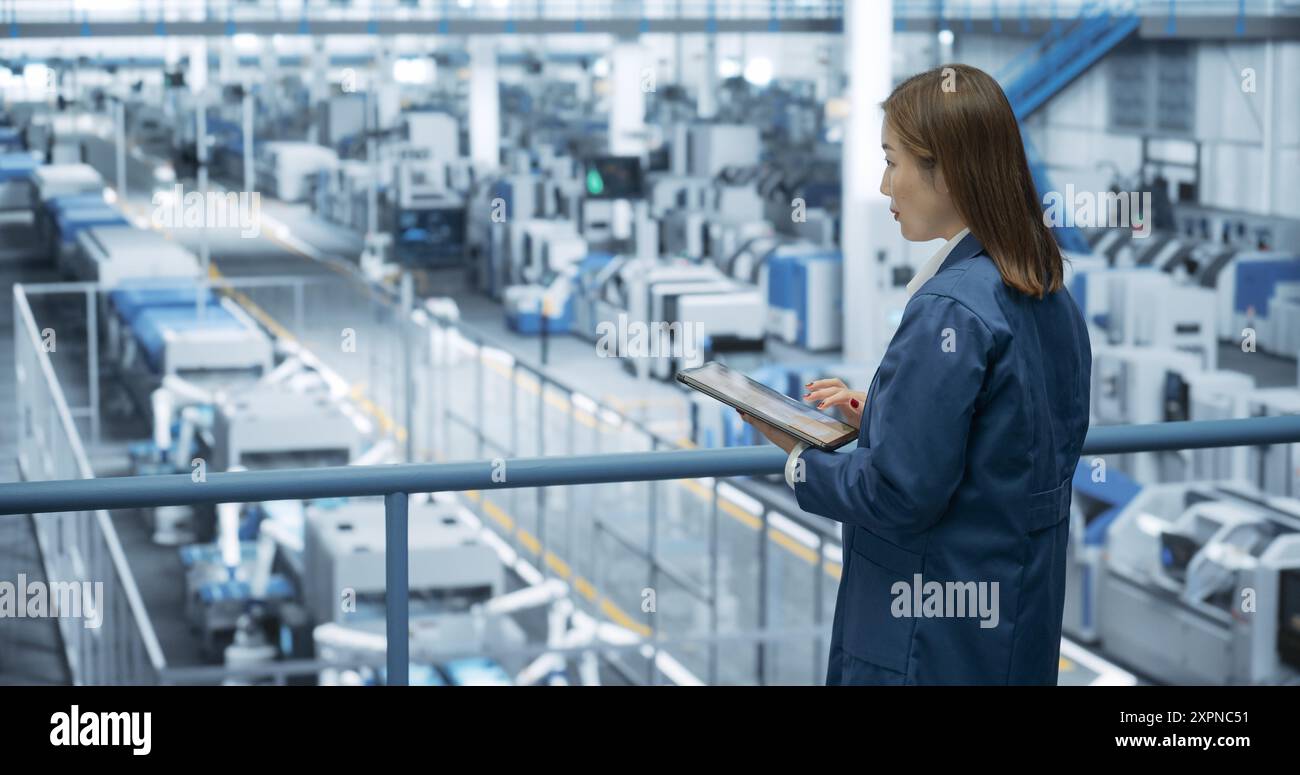 Junge asiatische Ingenieurin, die auf einer Plattform steht und einen Tablet-Computer in einer Elektronikfabrik benutzt. Maschinen werden gewartet, Spezialist überwacht den Fortschritt mithilfe der Online-Software Stockfoto