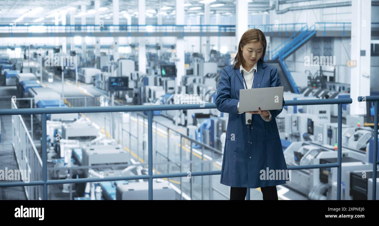 Asiatische Ingenieurin, die in einer Fabrik steht, an Laptopcomputern arbeitet, Technologien zur Herstellung von Elektronik in einer modernen automatisierten Fertigung mit Roboterarmen erforscht und entwickelt Stockfoto