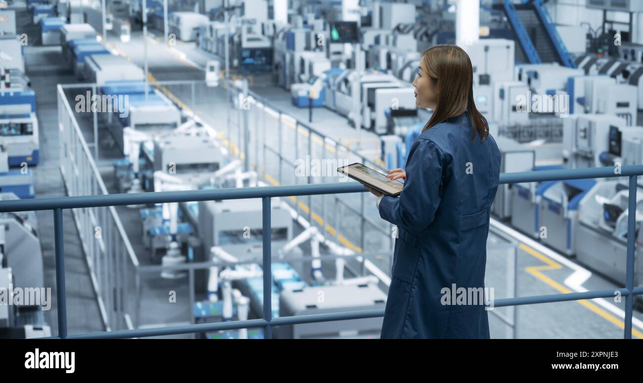 Junge asiatische Ingenieurin, die auf einer Plattform steht und einen Tablet-Computer in einer Elektronikfabrik benutzt. Maschinen werden gewartet, Spezialist überwacht den Fortschritt mithilfe der Online-Software Stockfoto
