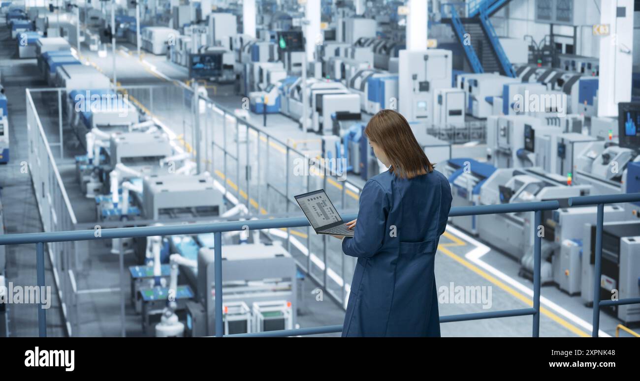 Asiatische Ingenieurin, die in einer Fabrik steht, an Laptopcomputern arbeitet, Technologien zur Herstellung von Elektronik in einer modernen automatisierten Fertigung mit Roboterarmen erforscht und entwickelt Stockfoto