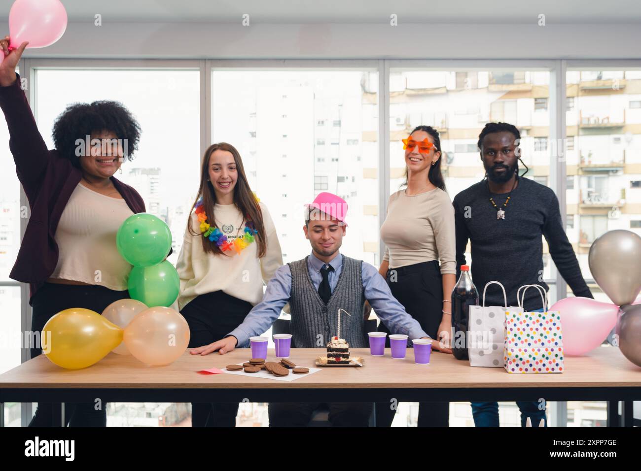 Kleine Versammlung von Bürokollegen, die einen der Kollegen zum Geburtstag feiern, Gruppe von multiethnischen Menschen mit Ballons, Geburtstagshüte und Stockfoto