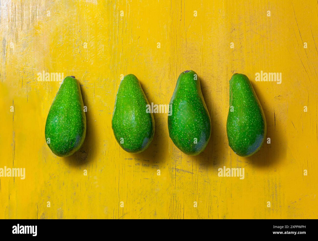 Vier grüne Avocados sind ordentlich in einer Reihe auf einer leuchtend gelben Holzoberfläche angeordnet Stockfoto