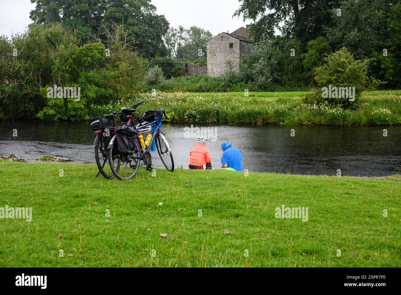 Grassington Yorkshire UK-27. Juli 2024. Radfahrer ruhen sich an einem ruhigen Fluss aus und genießen an einem bewölkten Tag die malerische Schönheit und die friedliche Umgebung. Stockfoto