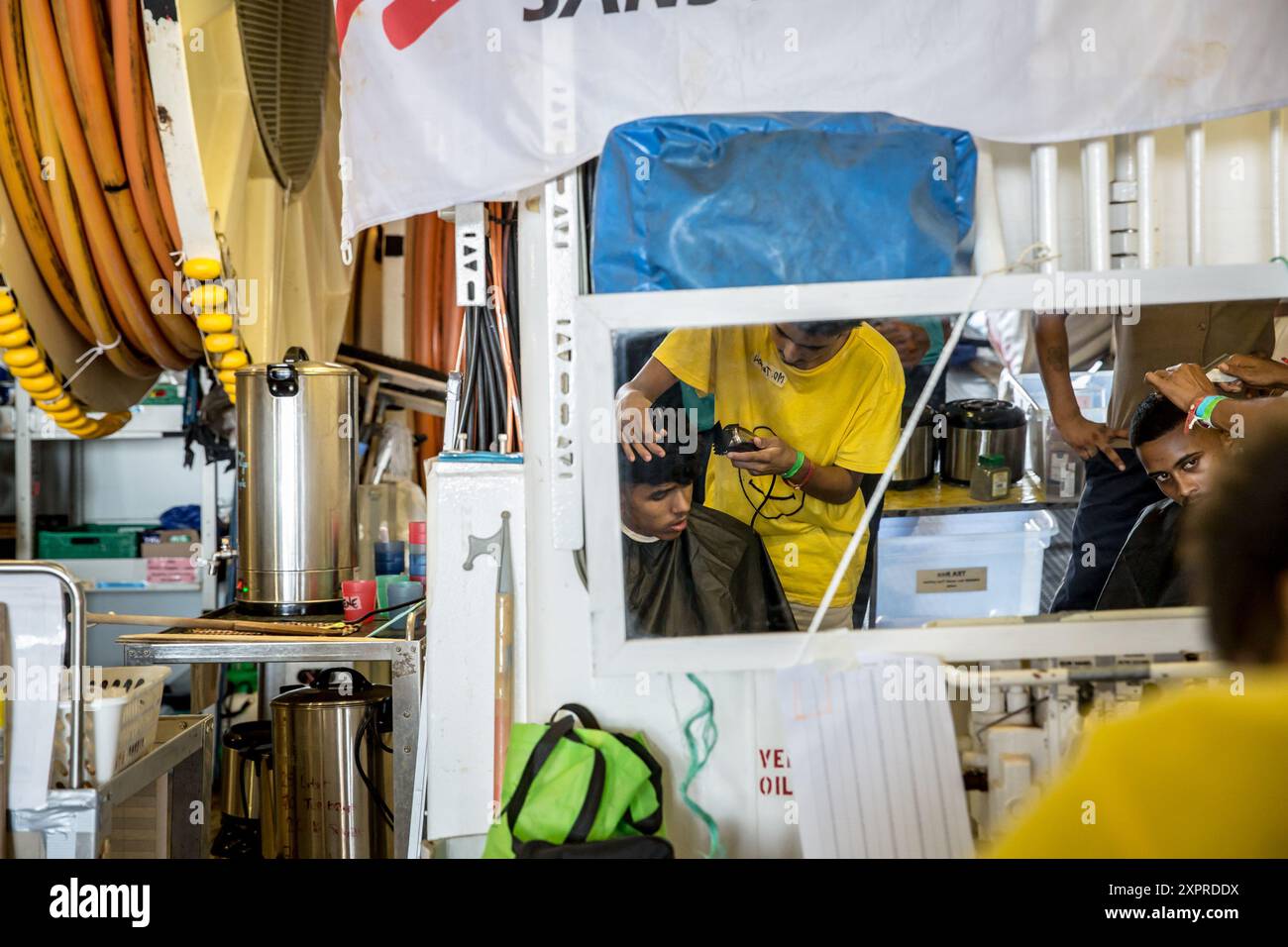 Auf See. August 2024. Bangladeshis, die von der Besatzung der Geo Barents gerettet wurden, einem Rettungsschiff, das im zentralen Mittelmeer von Médecins sans Frontières betrieben wird, geben einander Frisuren, nachdem ihnen Ausrüstung zur Verfügung gestellt wurde. Viele sind vor kurzem aus Schmugglern oder Haftanstalten geflohen. UN-Zahlen zeigen, dass seit 2014 mehr als 30.000 Menschen im Mittelmeer gestorben oder vermisst sind, während sie versucht haben, Europa zu erreichen. (Foto: Sally Hayden/SOPA Images/SIPA USA) Credit: SIPA USA/Alamy Live News Stockfoto