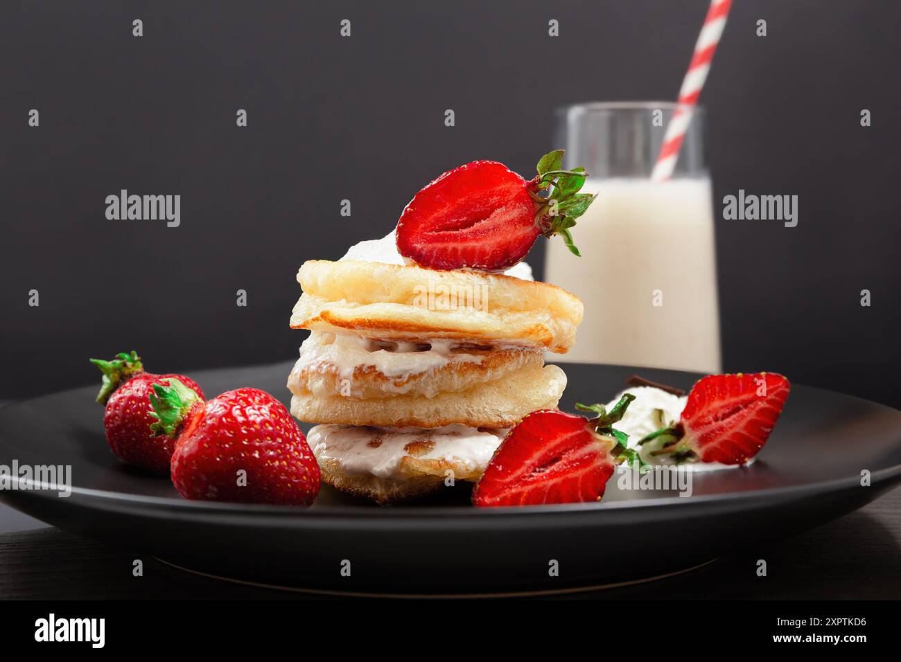 Ein Stapel Pfannkuchen mit Sahne und Erdbeeren auf einem schwarzen Teller, mit einem Glas Milchshake im Hintergrund Stockfoto