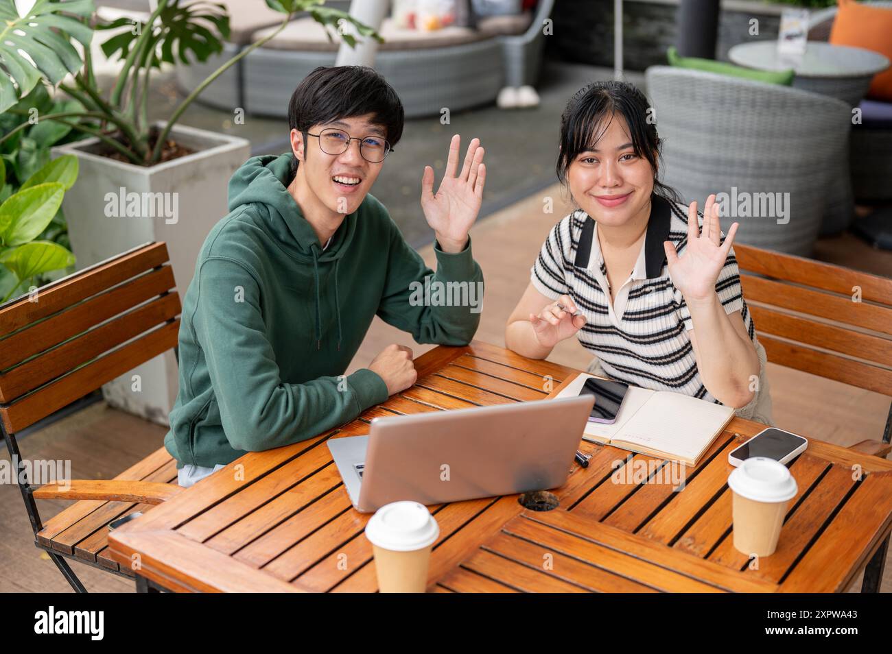 Zwei fröhliche, freundliche junge asiatische Freundinnen sitzen draußen an einem Tisch und winken mit der Hand zur Kamera. Freundschaft, Teamarbeit, Stockfoto