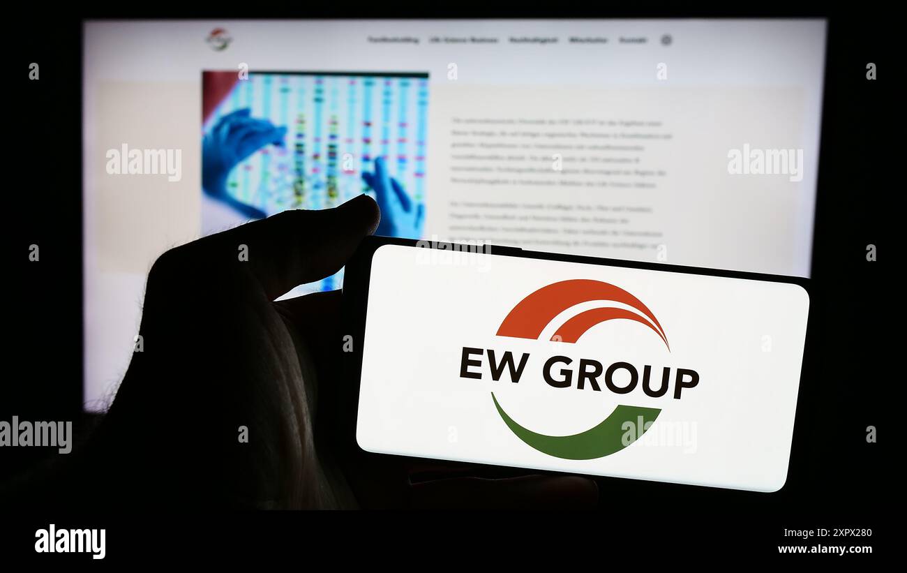 Person mit Mobiltelefon mit Logo des deutschen Tierzuchtunternehmens EW Group GmbH vor der Business-Webseite. Schwerpunkt auf dem Display des Telefons. Stockfoto