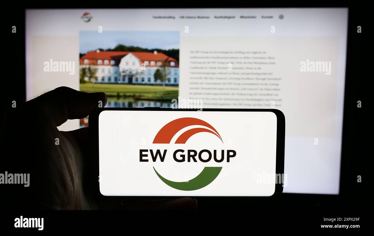 Person mit Handy mit Logo der deutschen Viehzuchtgesellschaft EW Group GmbH vor der Business-Website. Schwerpunkt auf dem Display des Telefons. Stockfoto
