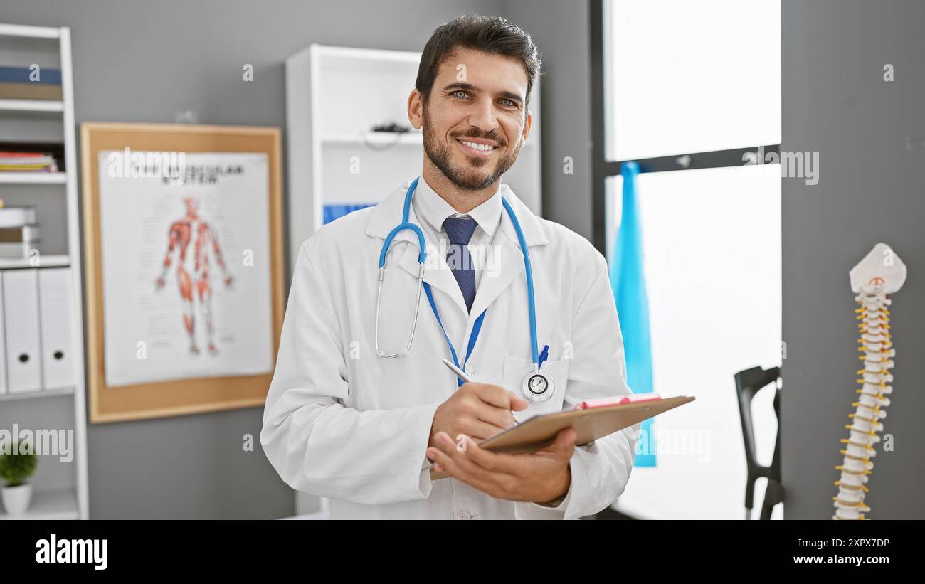 Gutaussehender hispanischer Mann im weißen Labormantel, der Notizen in einem Klinikinnenraum mit Muskel-Skelett-System-Chart macht. Stockfoto
