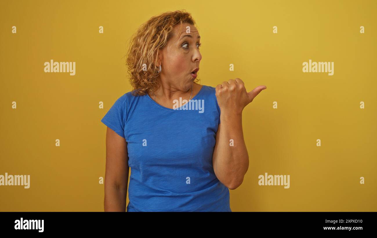 Eine hispanische Frau mittleren Alters in einem blauen Hemd gestet mit ihrem Daumen auf gelbem Hintergrund und vermittelt mit offenem Mund einen überraschten Ausdruck. Stockfoto