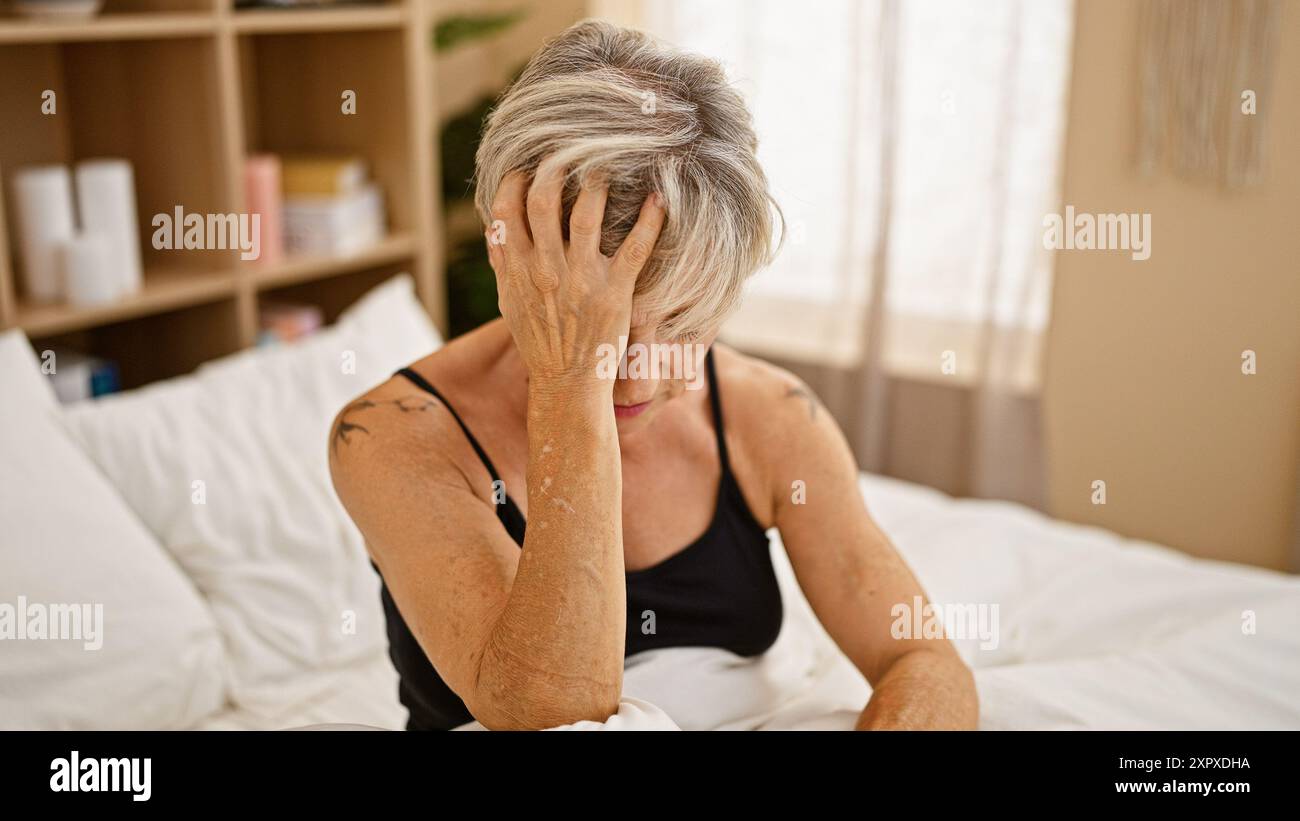 Reife Frau mit grauem Haar, die in einem Schlafzimmer gestresst aussieht und über persönliche Probleme in Innenräumen reflektiert. Stockfoto