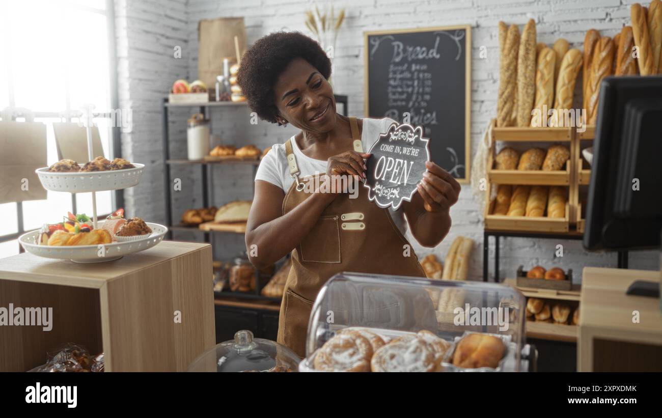 Frau mit lockigen Haaren lächelt, während sie das Schild in einer gemütlichen Bäckerei mit frischem Brot und Gebäck aufklappt, was ihren Stolz auf den Laden unterstreicht. Stockfoto