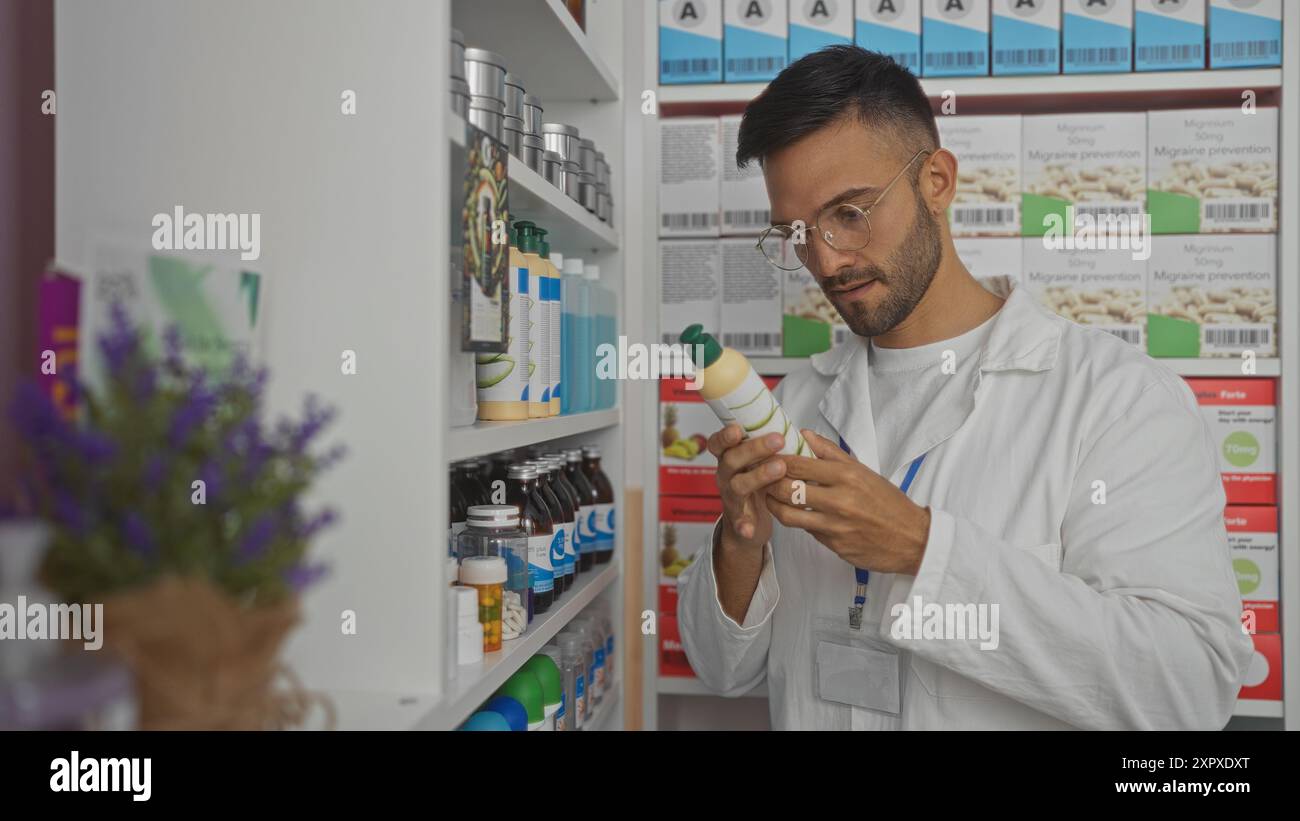 Ein junger hispanischer Mann in einer Apotheke, der einen weißen Laborkittel trägt und eine Produktflasche in einem Innenbereich voller Arzneiregale sorgfältig untersucht. Stockfoto