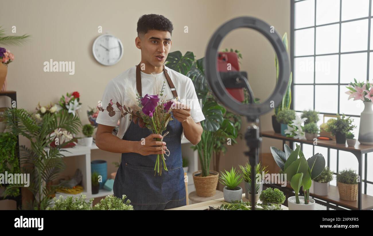 Ein junger Mann mit Schürze arrangiert Blumen in einem lichtdurchfluteten Blumenladen, umgeben von verschiedenen Pflanzen und einem Ringlicht. Stockfoto