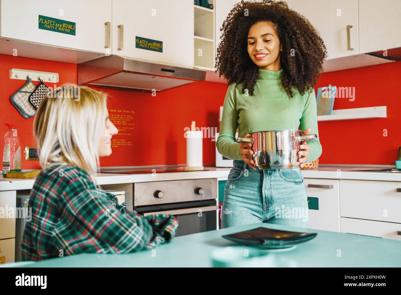 Zwei junge Frauen, eine mit lockigen Afro-Haaren und eine blonde, bereiten in einer leuchtend roten Küche Essen zu. Zeigt Vielfalt, Freundschaft und moderne Lebensweise Stockfoto