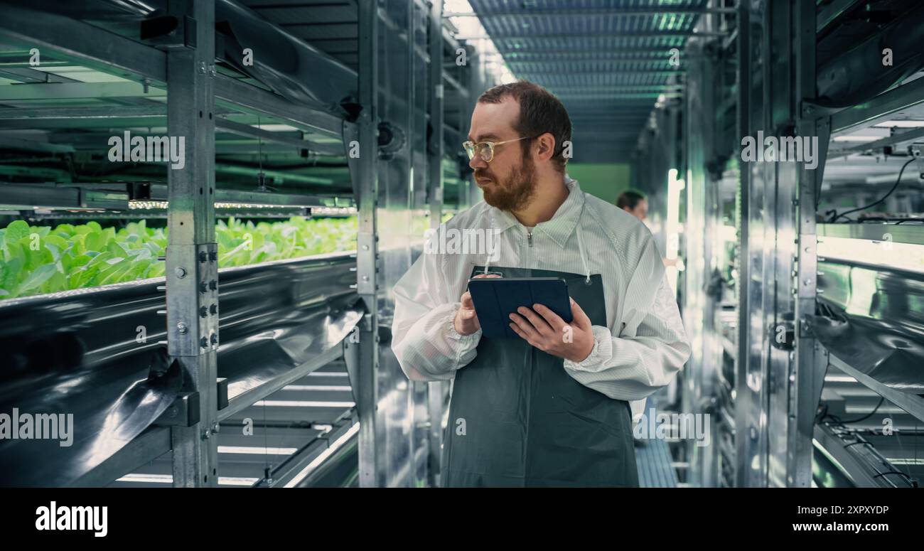 Biologiewissenschaftler, der in einer vertikalen Farm neben Rack mit natürlichen Öko-Pflanzen arbeitet. Landwirtschaftsingenieur mit Tablet-Computer, Organisieren und Analysieren von Erntegutinformationen vor der Verteilung Stockfoto