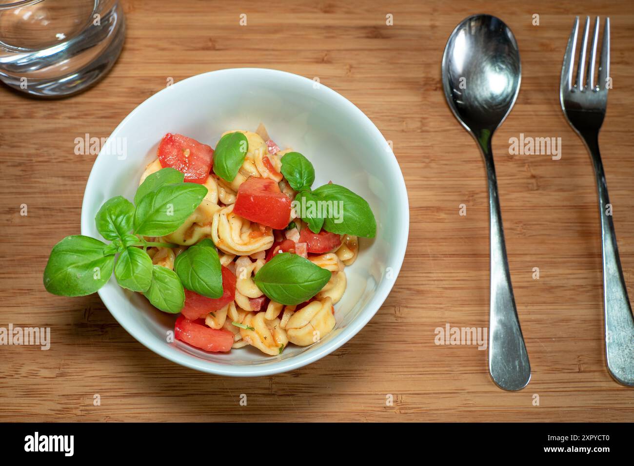 Tortellini-Salat in einer kleinen Schüssel mit Besteck und einem Glas Wasser auf einem Holztisch Stockfoto