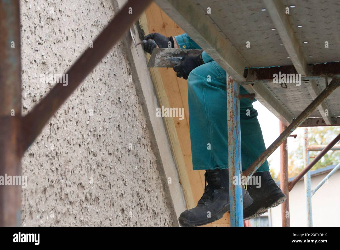Nahaufnahme der Beine eines Bauarbeiters, der mit Sicherheitsschuhen auf einem Gerüst sitzt und etwas auf dem Dachgiebel mit Kellen und ce repariert Stockfoto
