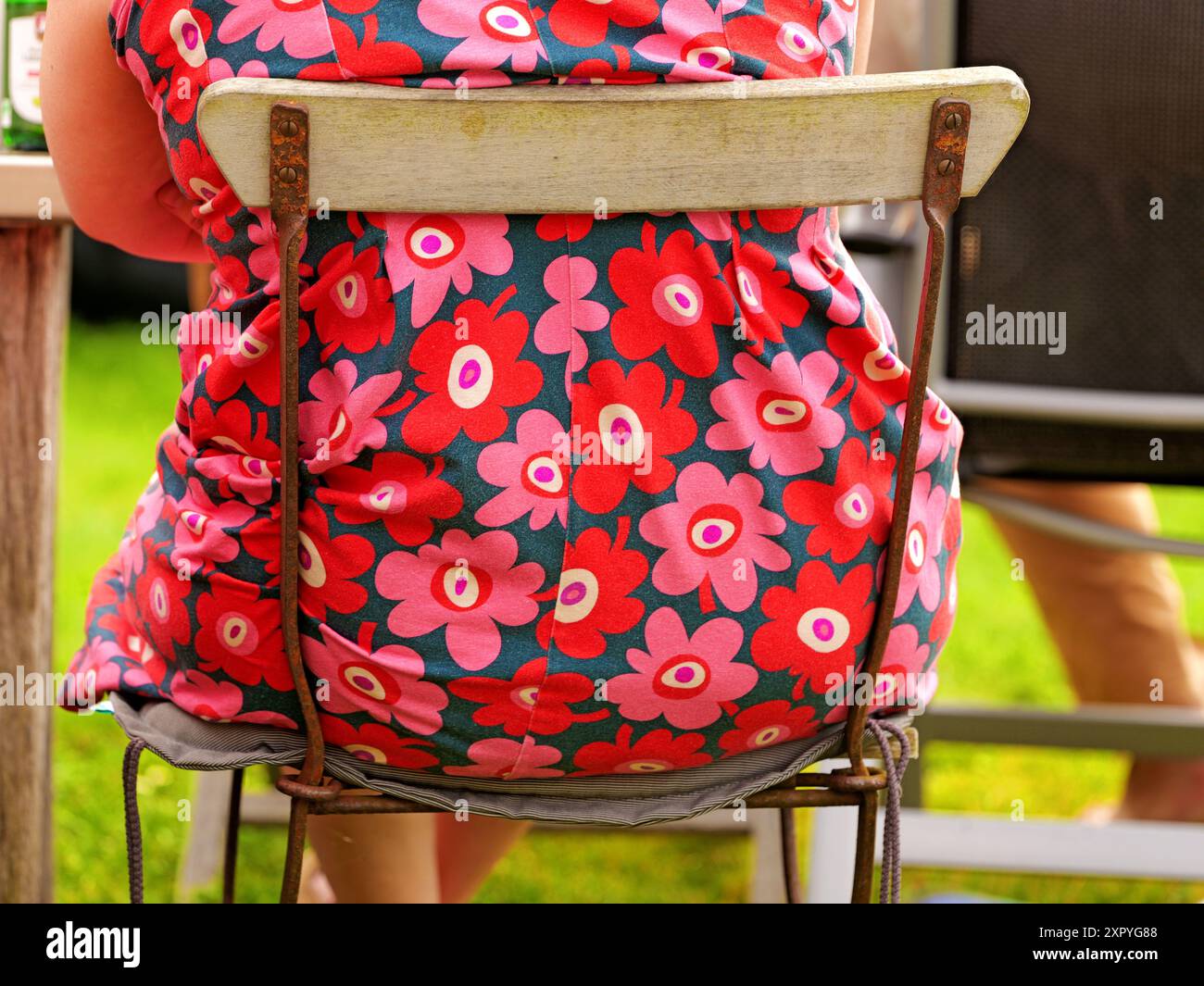 Frau in einem Retro-Blumenkleid auf einem alten Gartenstuhl. Die Kombination bringt ein Lächeln und ist perfekt als Textur und Hintergrund. Stockfoto