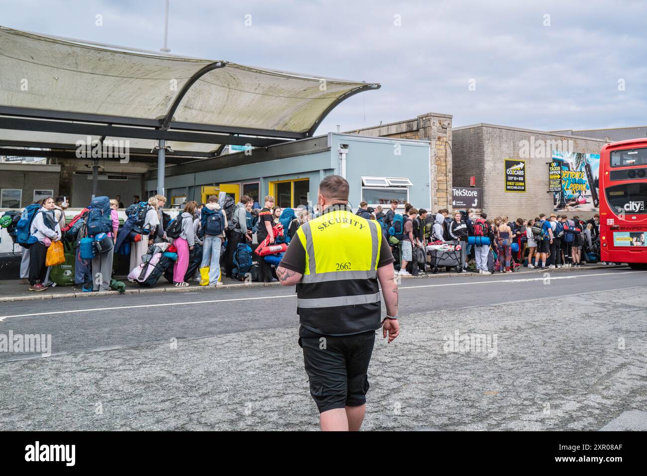 Ein Sicherheitsarbeiter überwacht junge Leute, die für Busse anstehen, um sie zum Boardmasters Festival Newquay in Cornwall in Großbritannien zu bringen. Stockfoto